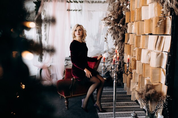 Hermosa mujer en vestido negro se sienta ante una pared de libros y decoración de Navidad