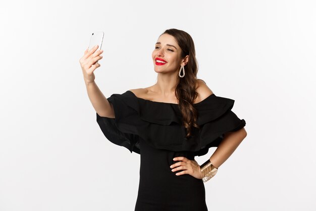 Hermosa mujer en vestido negro tomando selfie en fiesta, de pie sobre fondo blanco con smartphone.