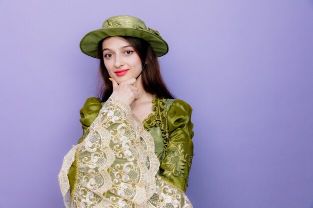 Hermosa mujer en vestido renacentista y sombrero con expresión pensativa sosteniendo la mano en la barbilla pensando en positivo en azul