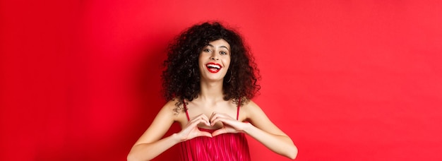 Hermosa mujer con vestido rojo de pelo rizado que muestra el símbolo del corazón y sonriente fondo de estudio feliz