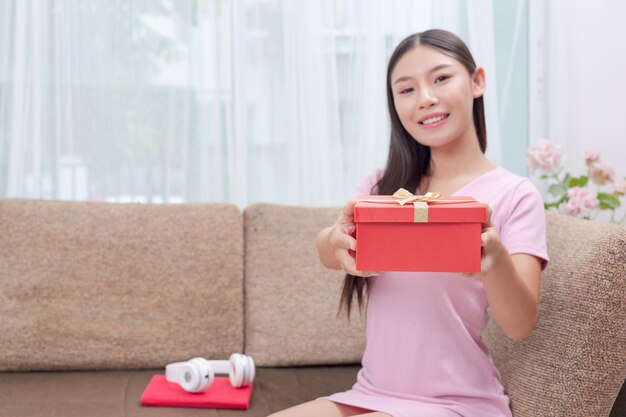 Hermosa mujer en vestido rosa, sentada en el sofá, abriendo una caja de regalo.