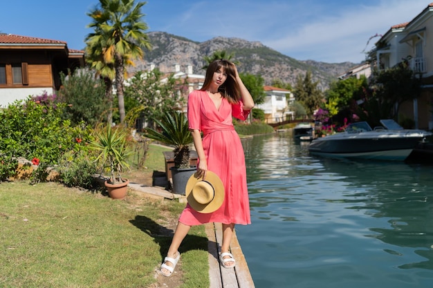 Hermosa mujer viajera con elegante vestido rosa y sombrero de paja posando sobre un complejo de lujo en la provincia de Gocek Mugla, Turquía