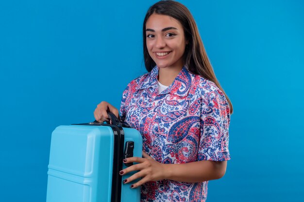 Hermosa mujer viajero sosteniendo maleta de viaje sonriendo alegremente positivo y feliz listo para vacaciones de pie sobre espacio azul aislado