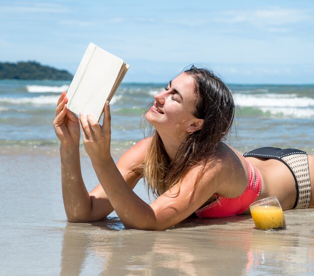 Hermosa niña acostada en la playa leyendo un libro con bebidas frescas de verano y frutas tropicales, vacaciones