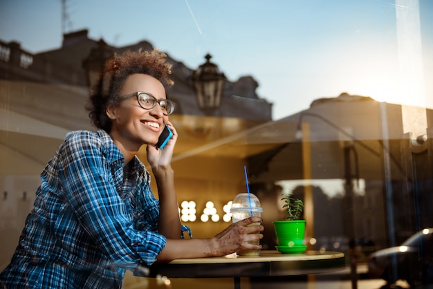 Hermosa niña africana sonriendo, hablando por teléfono, sentado en la cafetería. Disparo desde afuera.