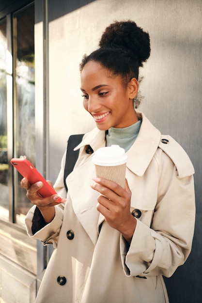 Hermosa niña afroamericana sonriente con elegante abrigo de trinchera con café para ir felizmente usando el celular al aire libre