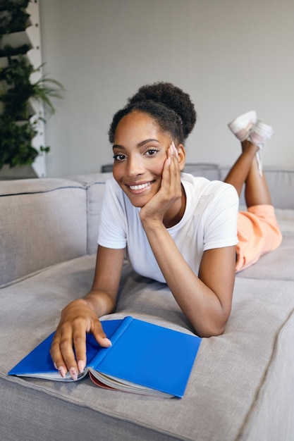 Hermosa niña afroamericana sonriente tendida en un sofá con un libro mirando felizmente a la cámara en una casa moderna