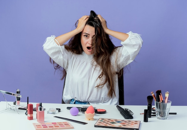 Hermosa niña ansiosa se sienta a la mesa con herramientas de maquillaje sostiene la cabeza mirando aislado en la pared púrpura