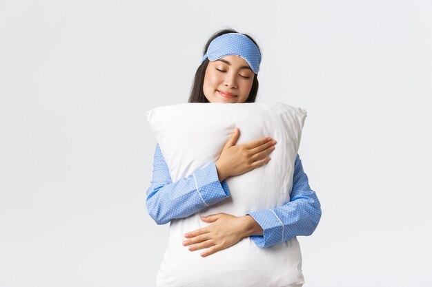 Hermosa niña asiática sonriente en máscara para dormir y pijama, teniendo un dulce sueño, abrazando la almohada con una sonrisa tonta y ojos cerrados, de pie sobre un fondo blanco, sin querer despertarse.