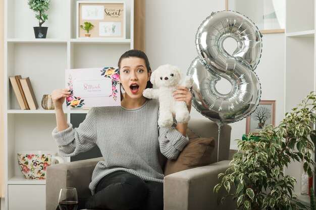 Hermosa niña asustada en el día de la mujer feliz con tarjeta de felicitación con oso de peluche sentado en un sillón en la sala de estar