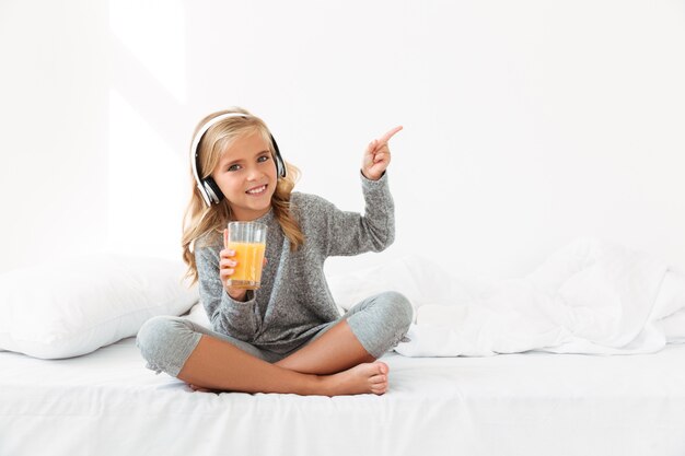 Hermosa niña en auriculares con vaso de jugo de naranja, apuntando con el dedo, mientras está sentado en la cama