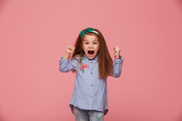 Hermosa niña en cabello aro y ropa de moda apretando los puños gritando de felicidad y admiración