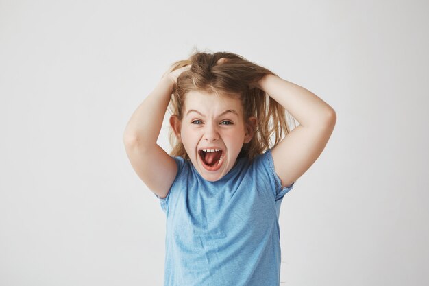 Hermosa niña con cabello claro gritando en voz alta, sosteniendo el cabello con las manos, asustada con una gran araña en la pared de la habitación.