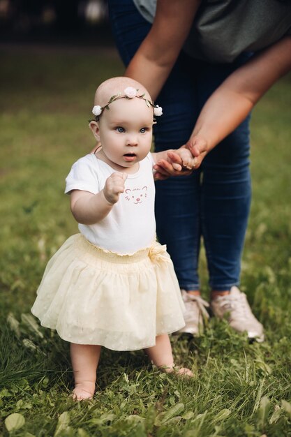 Hermosa niña con cabello corto y rubio y bonita sonrisa en vestido blanco se sienta en un césped en el parque en verano con su madre