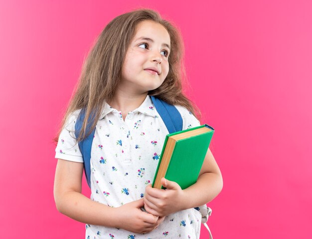 Hermosa niña con cabello largo con mochila sosteniendo portátil mirando a un lado con una sonrisa en la cara feliz de pie sobre la pared rosa