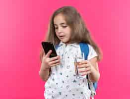 Foto gratuita hermosa niña con cabello largo con mochila sosteniendo smartphone y vaso de agua sonriendo confiado de pie sobre la pared rosa