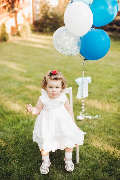 Hermosa niña caucásica con cabello rubio ondulado corto en vestido blanco se sienta en una silla en el jardín cerca de los globos