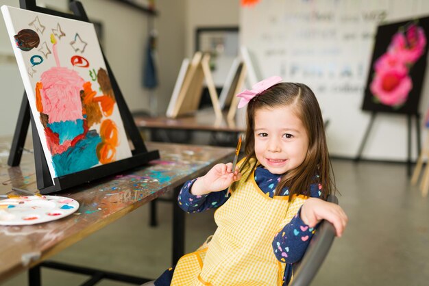 Hermosa niña caucásica con una expresión orgullosa sostiene un pincel y se sienta frente a su pintura terminada durante una clase de arte