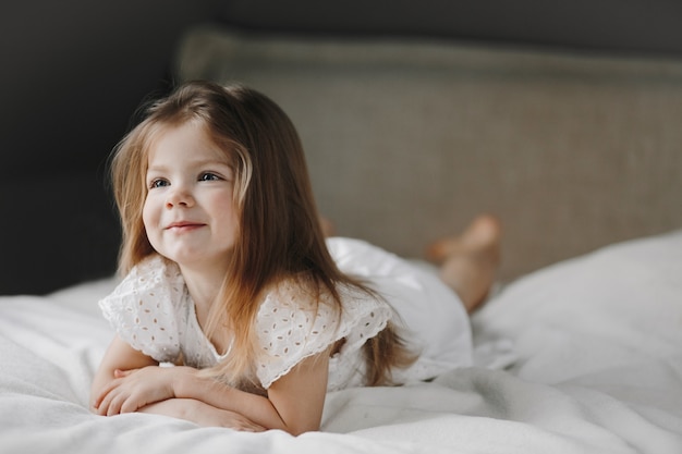 Hermosa niña caucásica pequeña está acostada en la cama blanca vestida con un vestido blanco y sonriendo, y mirando hacia un lado