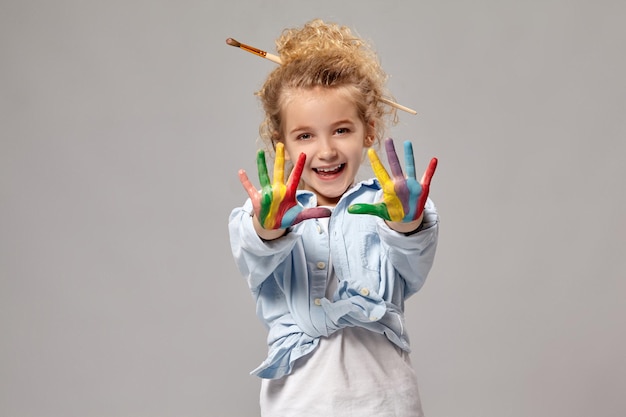 Hermosa niña con un cepillo en su elegante cabello rubio rizado, vestida con una camisa azul y una camiseta blanca. Ella está mostrando sus dedos pintados y mirando a la cámara, sobre un fondo gris.