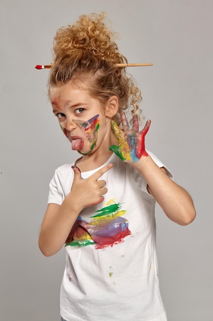 Hermosa niña con un cepillo en su hermoso corte de pelo, vestida con una camiseta manchada de blanco. Ella está posando con manos y mejillas pintadas, señalando su mano y mostrando una lengua, en una espalda gris