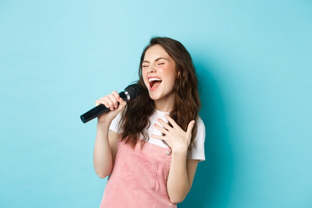 Hermosa niña despreocupada interpretar la canción, cantando en el micrófono con pasión, jugando karaoke, de pie sobre fondo azul.