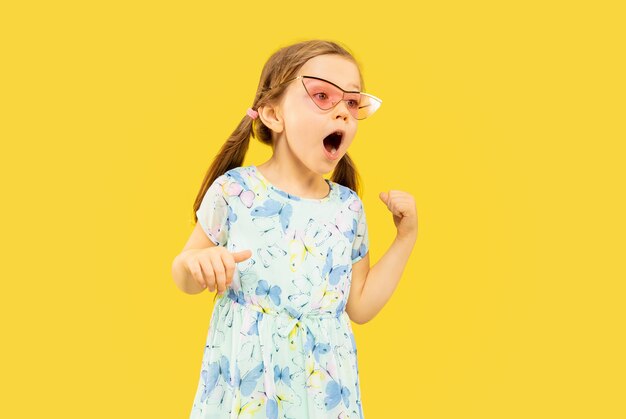 Hermosa niña emocional aislada en el espacio amarillo. Retrato de media longitud de niño feliz de pie y con un vestido y gafas de sol rojas