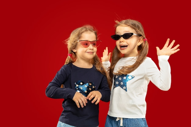 Foto gratuita hermosa niña emocional aislada en el espacio rojo. retrato de media longitud de hermanas felices o amigos con gafas de sol rojas y negras