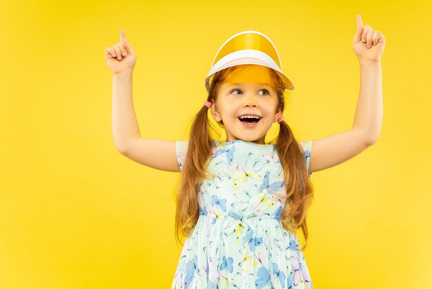 Hermosa niña emocional aislada. Retrato de niño feliz con vestido y gorra naranja apuntando hacia arriba. Concepto de verano, emociones humanas, infancia.