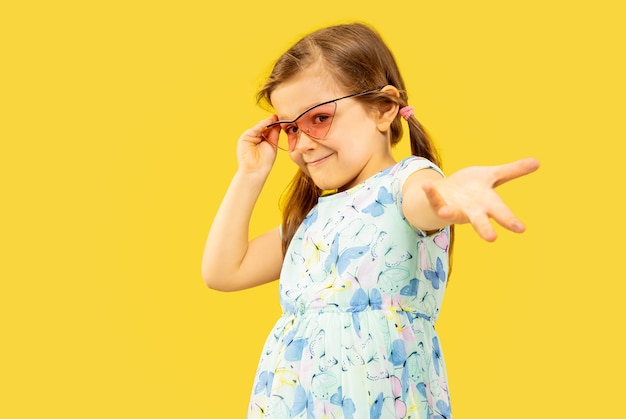 Hermosa niña emocional aislada sobre fondo amarillo. Retrato de medio cuerpo de un niño feliz de pie y con un vestido y gafas de sol rojas. Concepto de verano, emociones humanas, infancia.