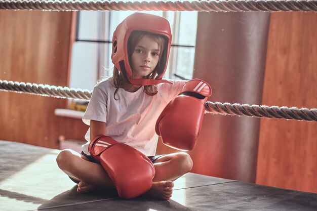 Hermosa niña está sentada en el ring de boxeo con uniforme de boxeador, guantes y casco.