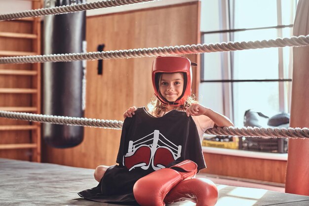 Hermosa niña está sentada en el ring de boxeo con uniforme de boxeador, guantes y casco.