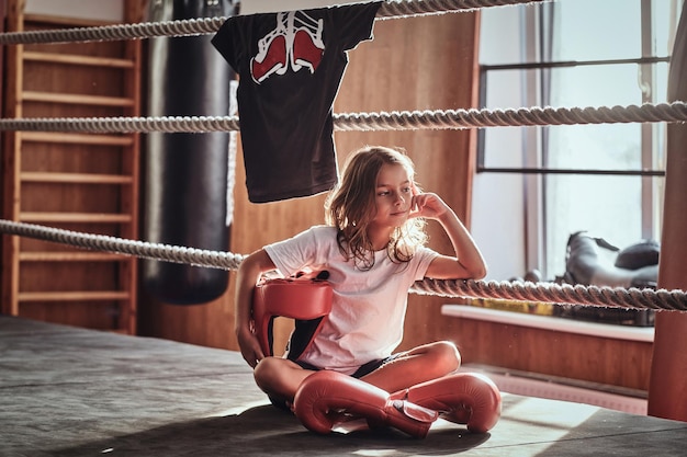 Hermosa niña está sentada en el ring de boxeo con uniforme de boxeador, guantes y casco.