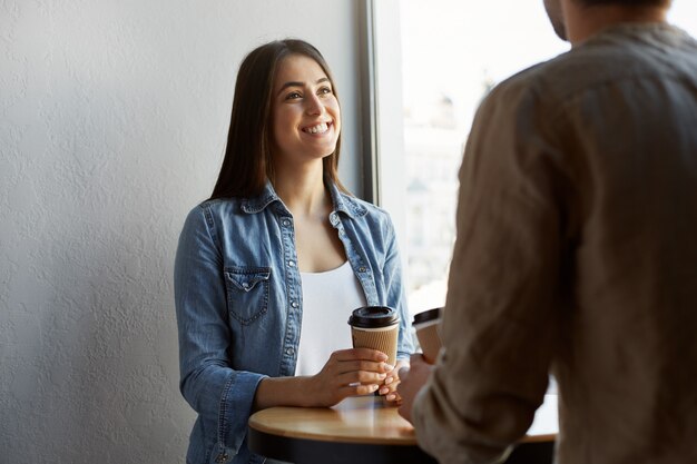 Hermosa niña feliz con el pelo oscuro en camiseta blanca debajo de la camisa vaquera, bebe café y sonriendo, escuchando la historia de un amigo de la fiesta de ayer.