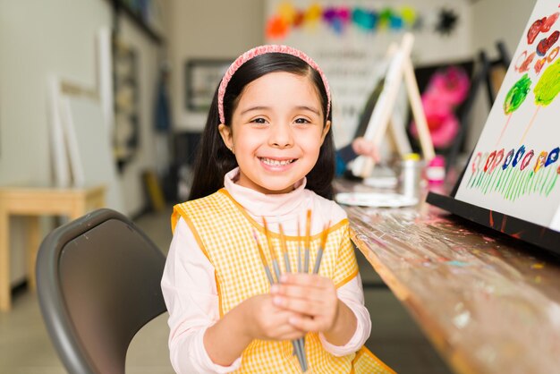 Hermosa niña hispana de primaria con un delantal que muestra sus pinceles que está usando en su clase de pintura en la escuela de arte