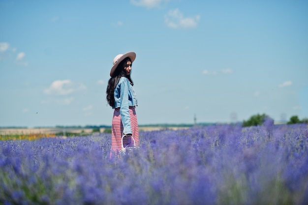 Hermosa niña india en vestido de verano y chaqueta de jeans en campo de lavanda púrpura con canasta en mano y sombrero