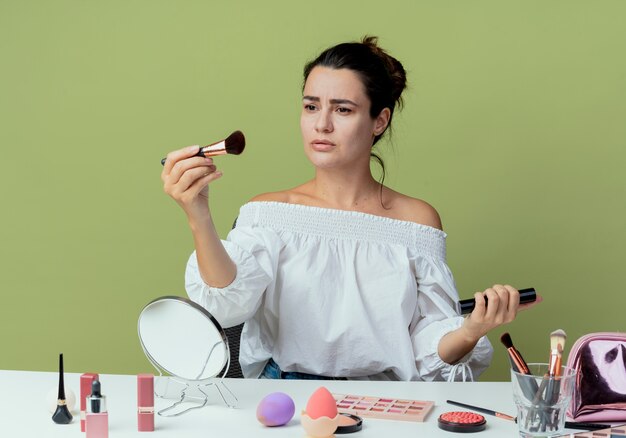 Hermosa niña molesta se sienta a la mesa con herramientas de maquillaje con cepillo y rímel aislado en la pared verde