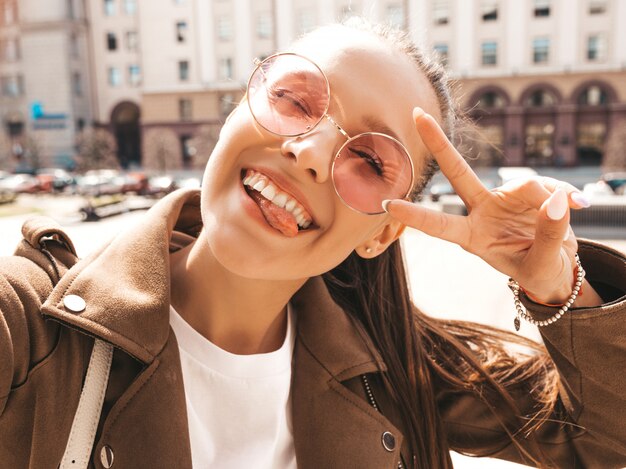 Hermosa niña morena sonriente en la chaqueta hipster de verano. Modelo tomando selfie en teléfono inteligente.