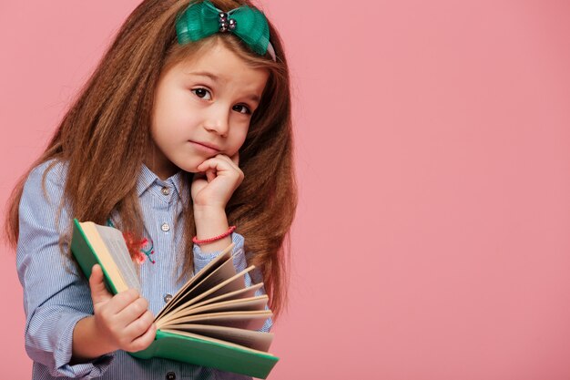 Hermosa niña pensativa con cabello largo y castaño apoyando la cabeza con la mano mientras lee un libro o aprende información