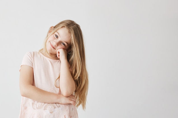 Hermosa niña pequeña con ojos azules y cabello claro en lindo vestido rosa sonríe y sostiene la mejilla con la mano.