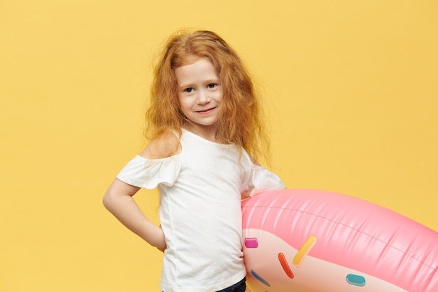 Hermosa niña posando aislada sosteniendo un tubo inflable rosa bajo el brazo yendo a la playa, con expresión facial feliz