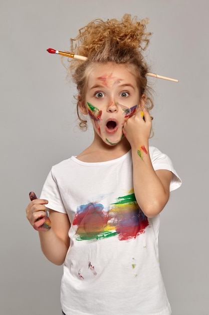 Hermosa niña que tiene un cepillo en su elegante cabello rubio rizado, vestida con una camiseta blanca, con los dedos pintados, se ve maravillada en un fondo gris.