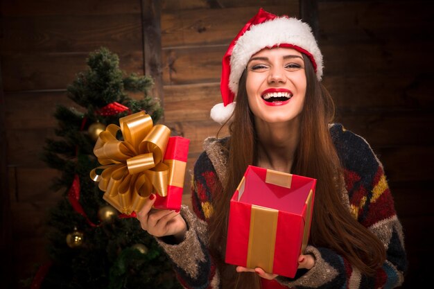 Hermosa niña riendo y abriendo un regalo de Papá Noel cerca del árbol de año nuevo. Señora feliz con labios rojos dentudos sonriendo en casa.