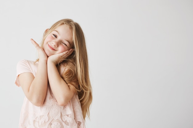 Hermosa niña rubia sonríe guiñando un ojo, posando, tocando la cara con sus manos en lindo vestido rosa. Niño feliz y encantado. Copia espacio