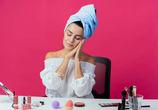 Foto gratuita hermosa niña soñolienta envuelto en una toalla para el cabello se sienta a la mesa con herramientas de maquillaje tiene las manos juntas cerca de la cara aislada en la pared rosa