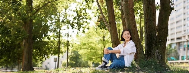 Foto gratuita hermosa niña sonriente se sienta cerca de un árbol en el parque disfrutando de la naturaleza al aire libre relajándose y descansando sobre productos frescos