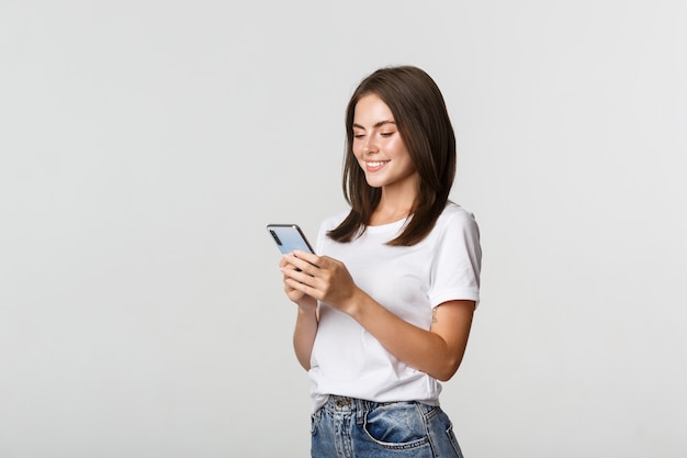 Hermosa niña sonriente con teléfono móvil, mirando smartphone satisfecho.