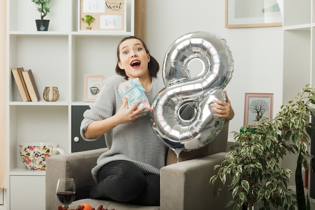 Foto gratuita hermosa niña sorprendida en el día de la mujer feliz sosteniendo el globo número ocho con presente sentado en un sillón en la sala de estar