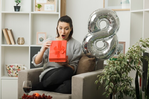 Hermosa niña sorprendida en el día de la mujer feliz sosteniendo y mirando al presente sentado en un sillón en la sala de estar