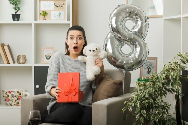 Hermosa niña sorprendida en el día de la mujer feliz sosteniendo presente con oso de peluche sentado en un sillón en la sala de estar